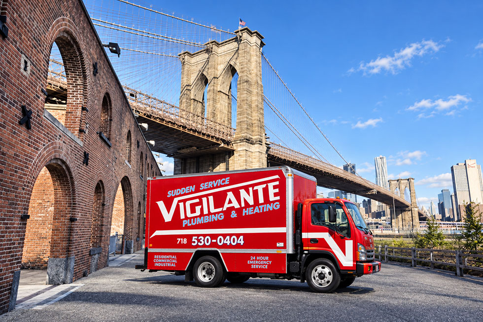 Red Vigilante Plumbing and Heating service truck parked near the Brooklyn Bridge beside a historic brick building.